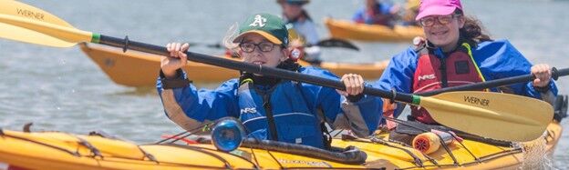 The image shows two people kayaking on a sunny day. They are both wearing life jackets and paddling with yellow paddles. The kayaks are yellow, and there are other kayaks in the background. The person in the front is wearing a green hat and glasses, while the person in the back is wearing a pink hat.